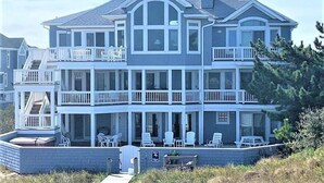 A two-story house with a balcony on the first floor and a covered patio area with outdoor seating. - Oceanfront, Heated Pool, Elevator, Book today! (Corolla)