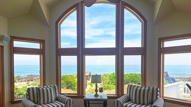 A room with large arched windows offering a view of the ocean. There are two striped armchairs and a small table with a lamp. - Oceanfront, Heated Pool, Elevator, Book today! (Corolla)