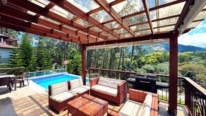 A covered outdoor area with a pool, wicker furniture, and a view of a forested landscape. - Vista del Campo Country Club & Villas (Jarabacoa)