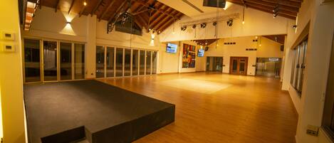 A spacious indoor area with wooden floors, a stage, and a high ceiling with exposed beams.