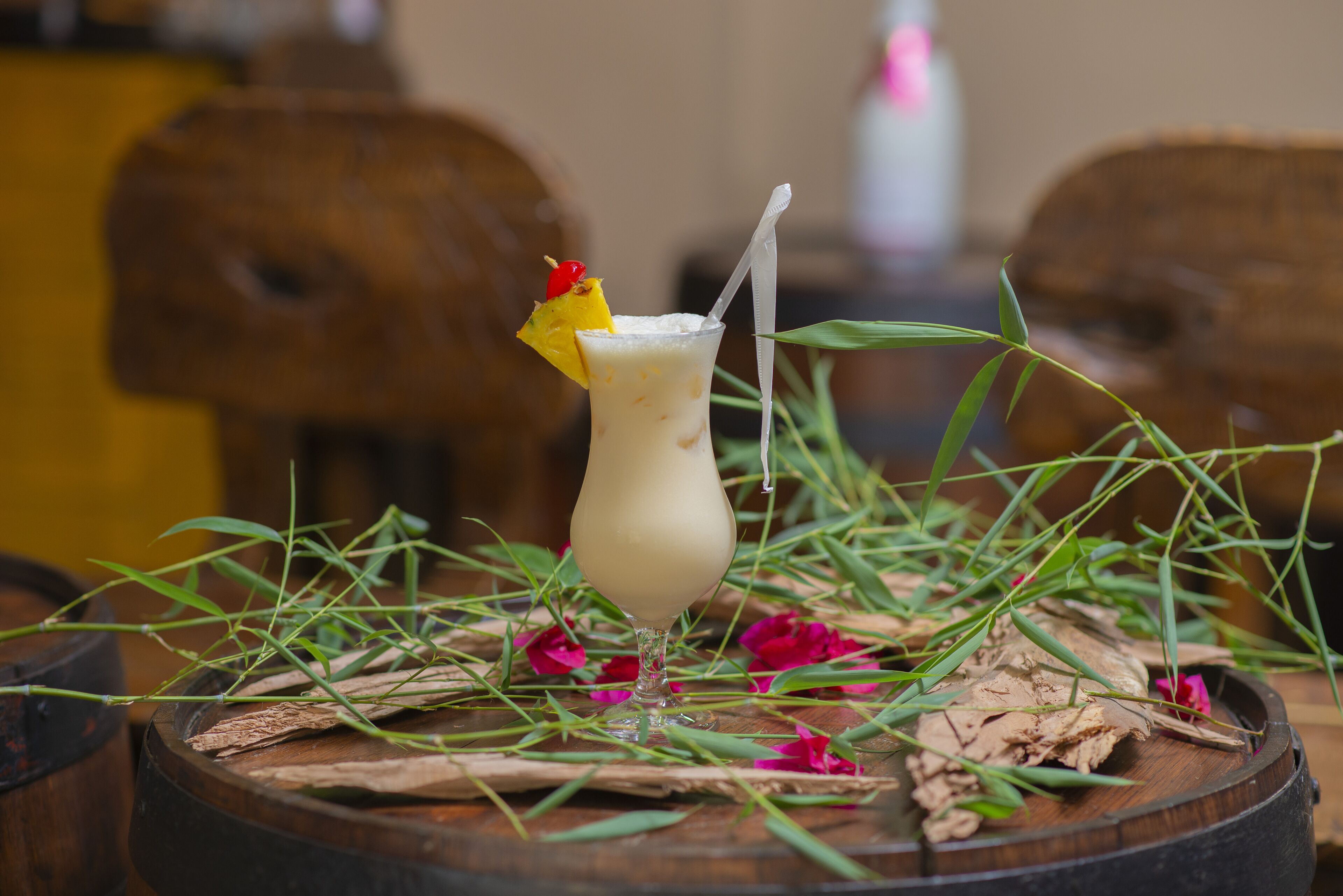A cocktail on a wooden barrel with green leaves and pink flowers around it.