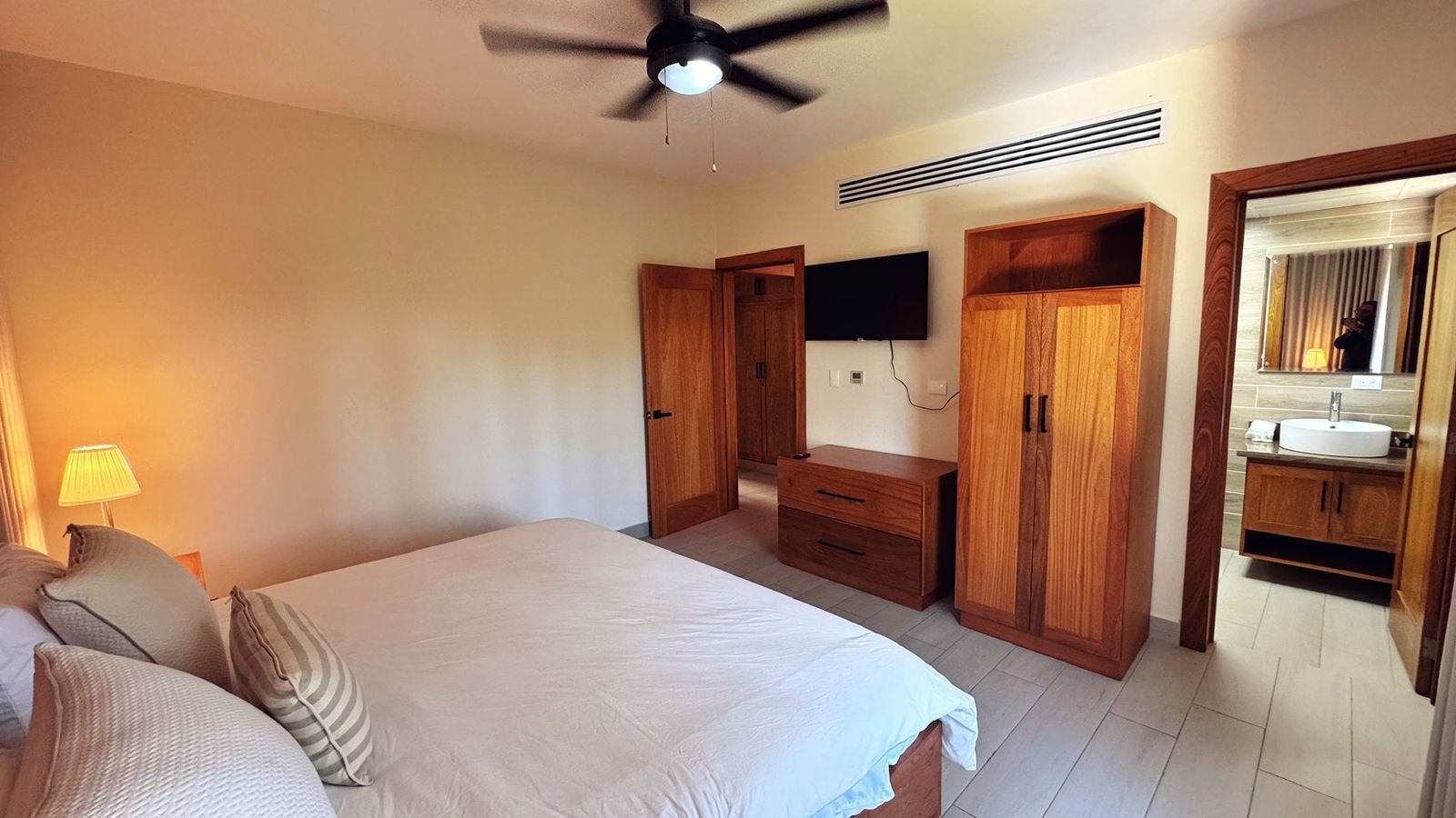 A bedroom with a bed, a nightstand, a desk, a wardrobe, a television, and a bathroom with a sink and mirror.