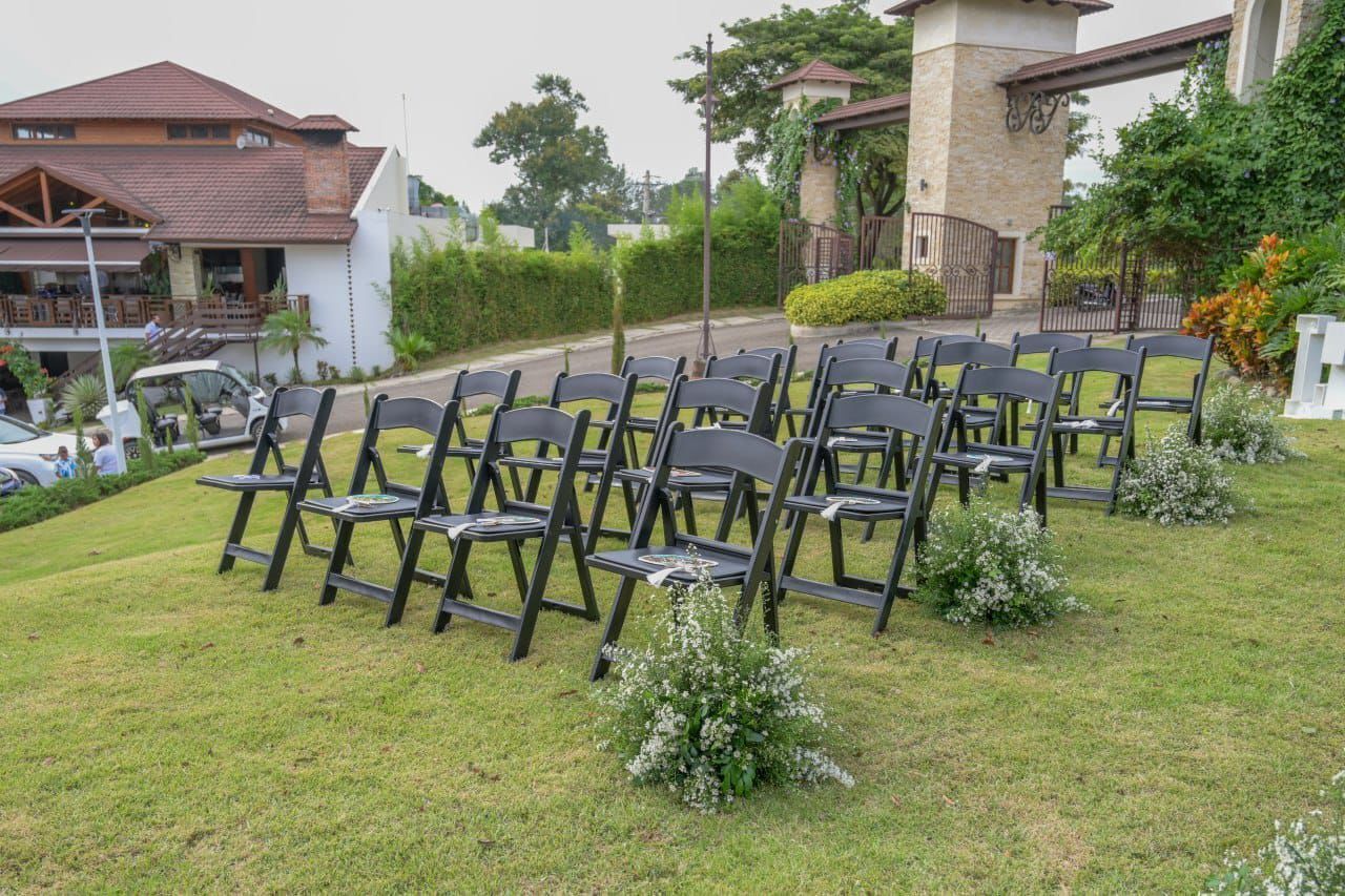 Filas de sillas negras dispuestas sobre un césped con pequeños arreglos florales blancos.