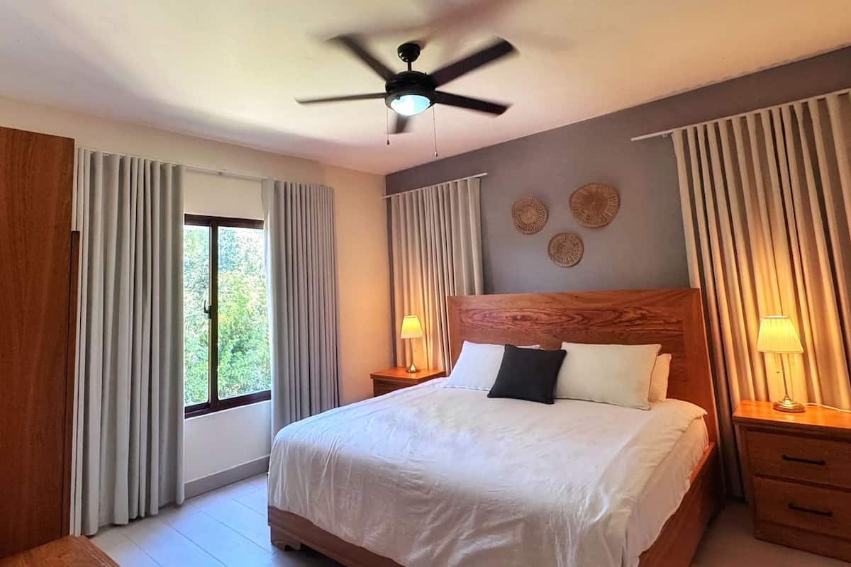 A bedroom with a bed, two bedside tables with lamps, a ceiling fan, and curtains.