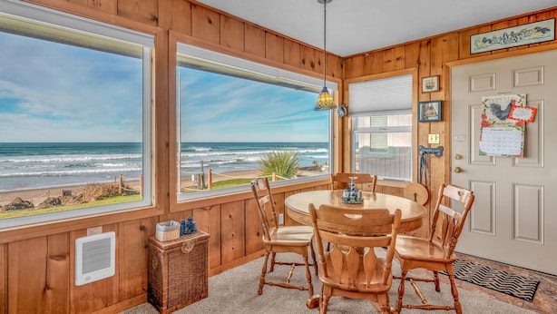 Una habitación con paneles de madera y un amplio ventanal con vista al océano, una mesa redonda de madera con sillas, y una puerta decorada con un calendario y un adorno de gallo.
