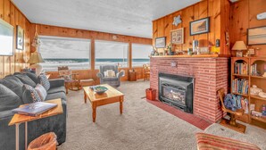 Una sala de estar acogedora con chimenea, paredes de madera, un ventanal con vista al océano, una estantería y una zona de estar confortable.