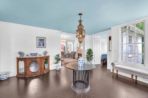 Un salón espacioso con una mesa redonda de cristal, una consola de madera con adornos azules y blancos, una lámpara colgante ornamentada y un ventanal con vistas al exterior.