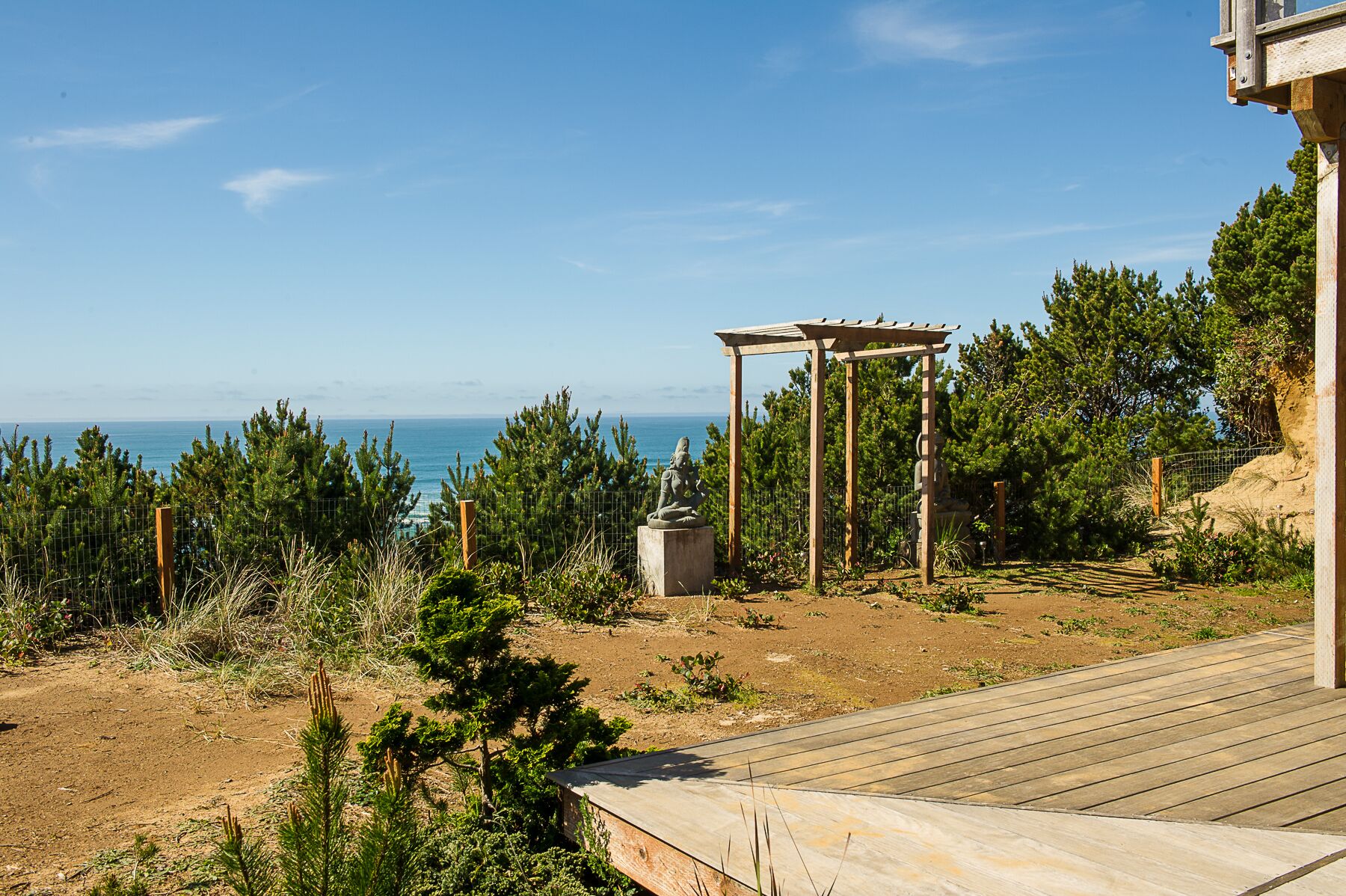 Een houten terras leidt naar een pergola met een standbeeld, met uitzicht op een kustlandschap met bomen en een heldere lucht.