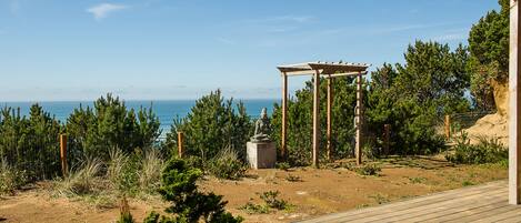 A wooden deck leading to a pergola with a statue, overlooking a coastal landscape with trees and a clear sky.