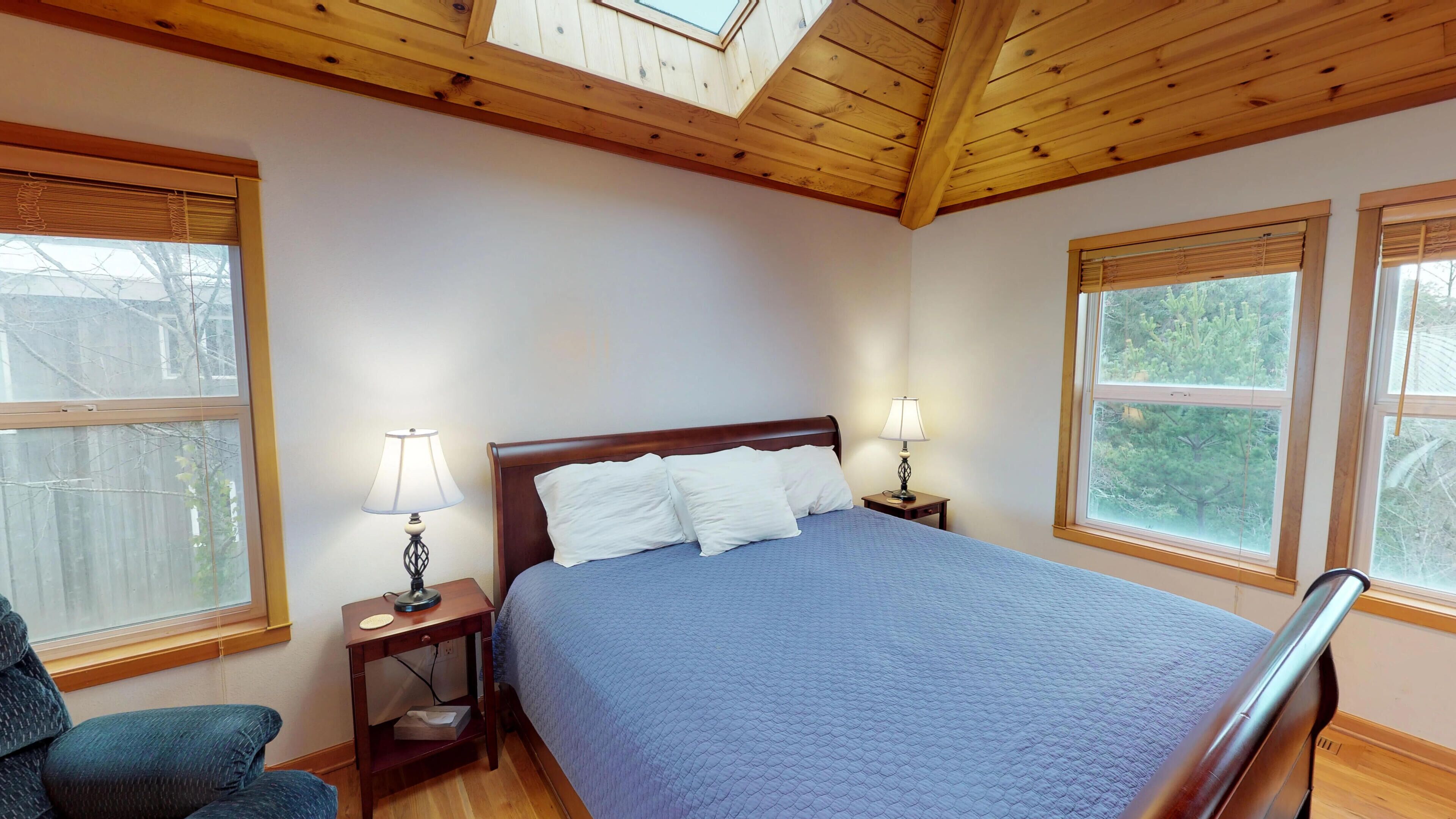 A bedroom with a wooden ceiling, a bed with white pillows, two bedside lamps, and large windows with blinds.