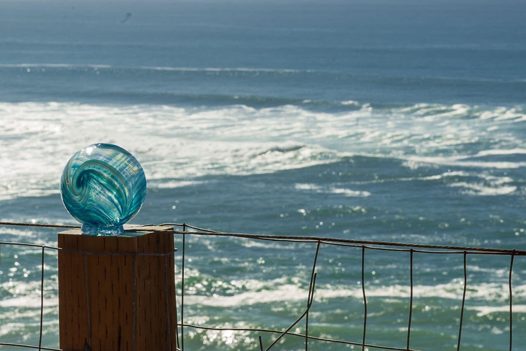 Una escultura de vidrio sobre un poste de madera, junto a una barandilla con vista al océano.