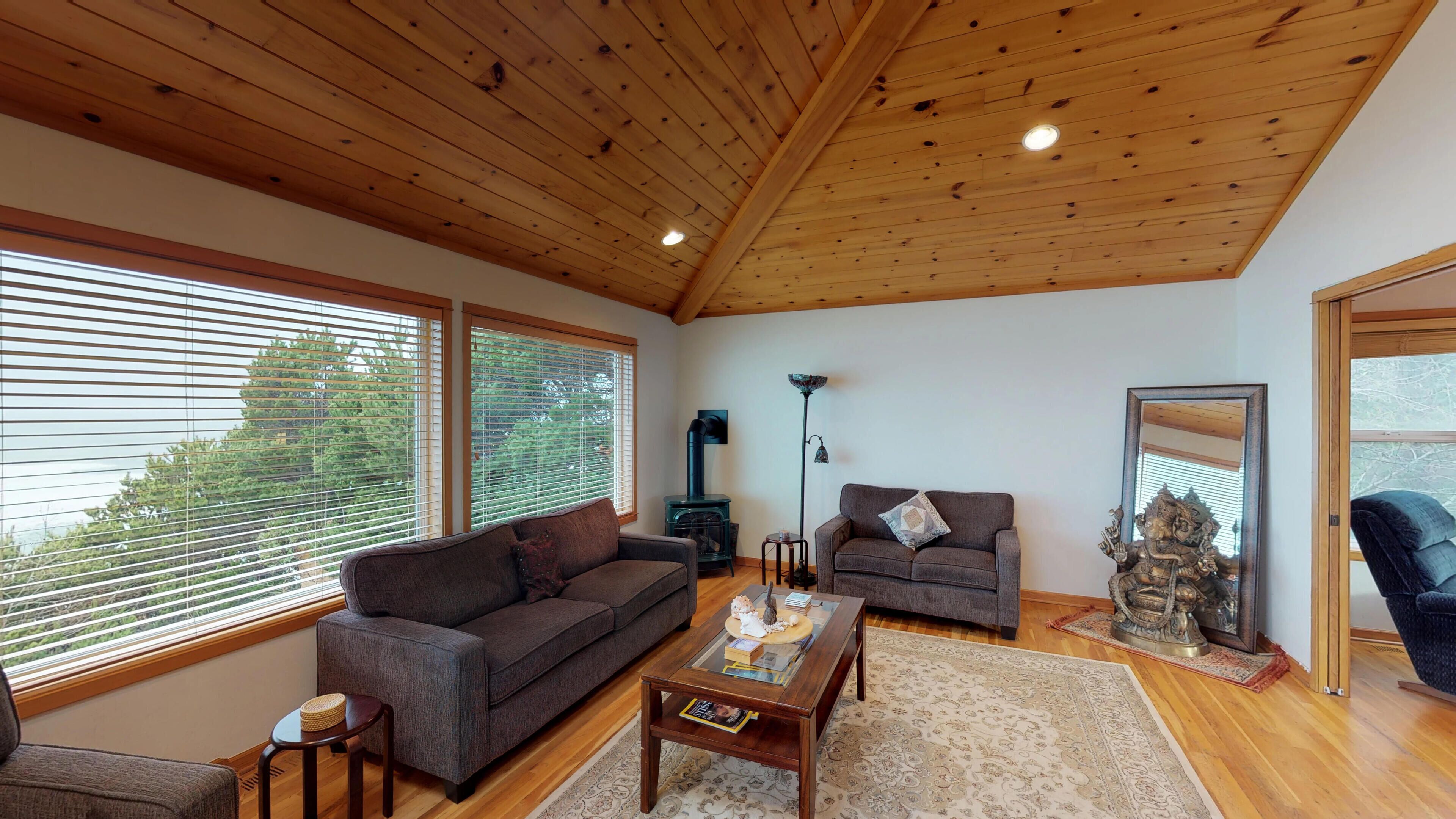 A spacious living room with a wooden ceiling, large windows, a sofa, a coffee table, and a fireplace.