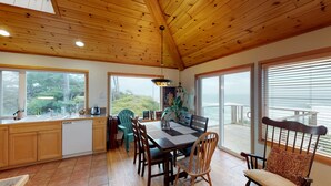 Een houten interieur met een eettafel, stoelen en uitzicht op de oceaan door grote ramen.