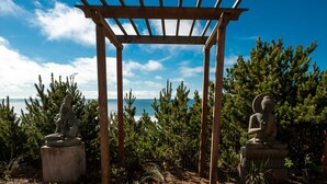 Een houten pergola met aan weerszijden twee stenen Boeddhabeelden, voor een achtergrond van altijdgroene bomen en een heldere lucht.
