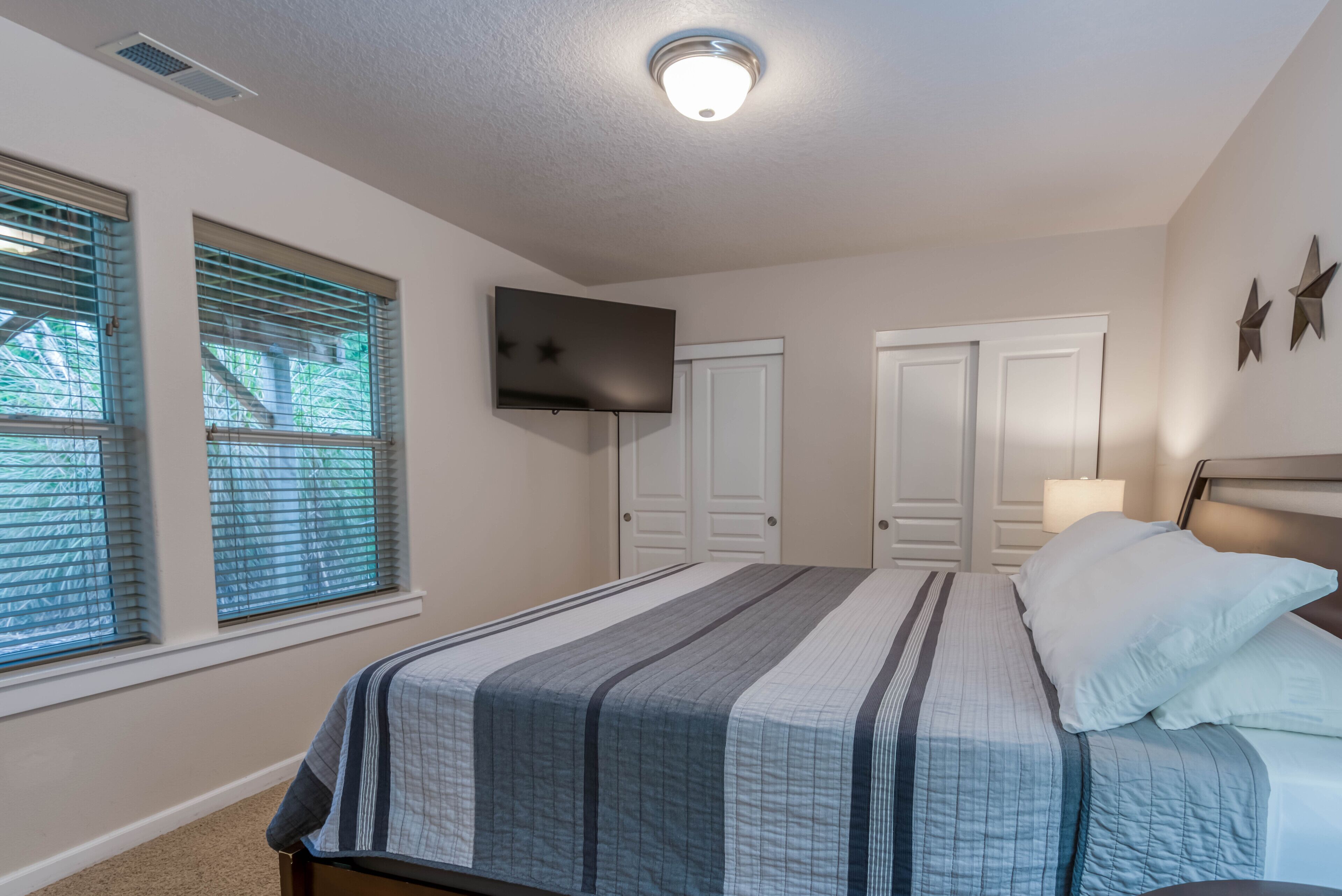 A bedroom with a bed, two windows, a TV, and a ceiling light.