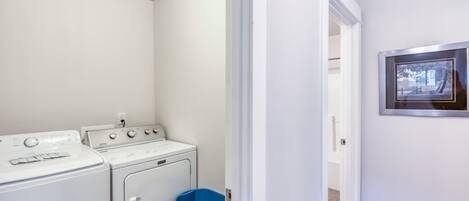 Una sala de bugaderia amb rentadora i assecadora, una cistella de roba blava i un quadre al mur.