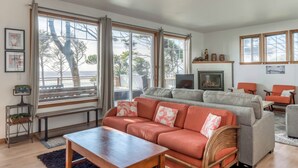 Una sala de estar con sofá, mesa de centro y chimenea. Hay ventanas amplias con cortinas, un televisor y una estantería.