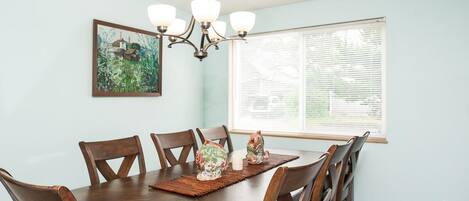 Une salle à manger avec une table en bois, quatre chaises, un lustre, une fenêtre avec des stores et un tableau au mur.