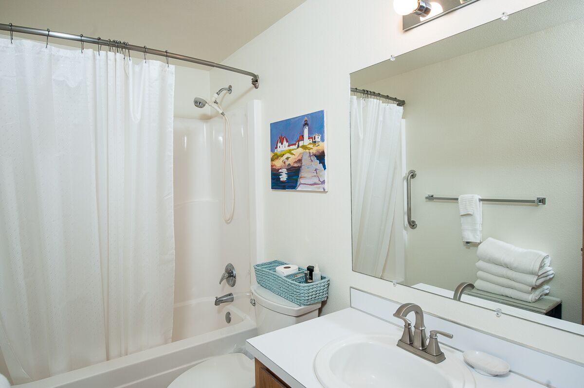 Un baño con ducha, lavamanos, espejo y un cuadro de un faro.