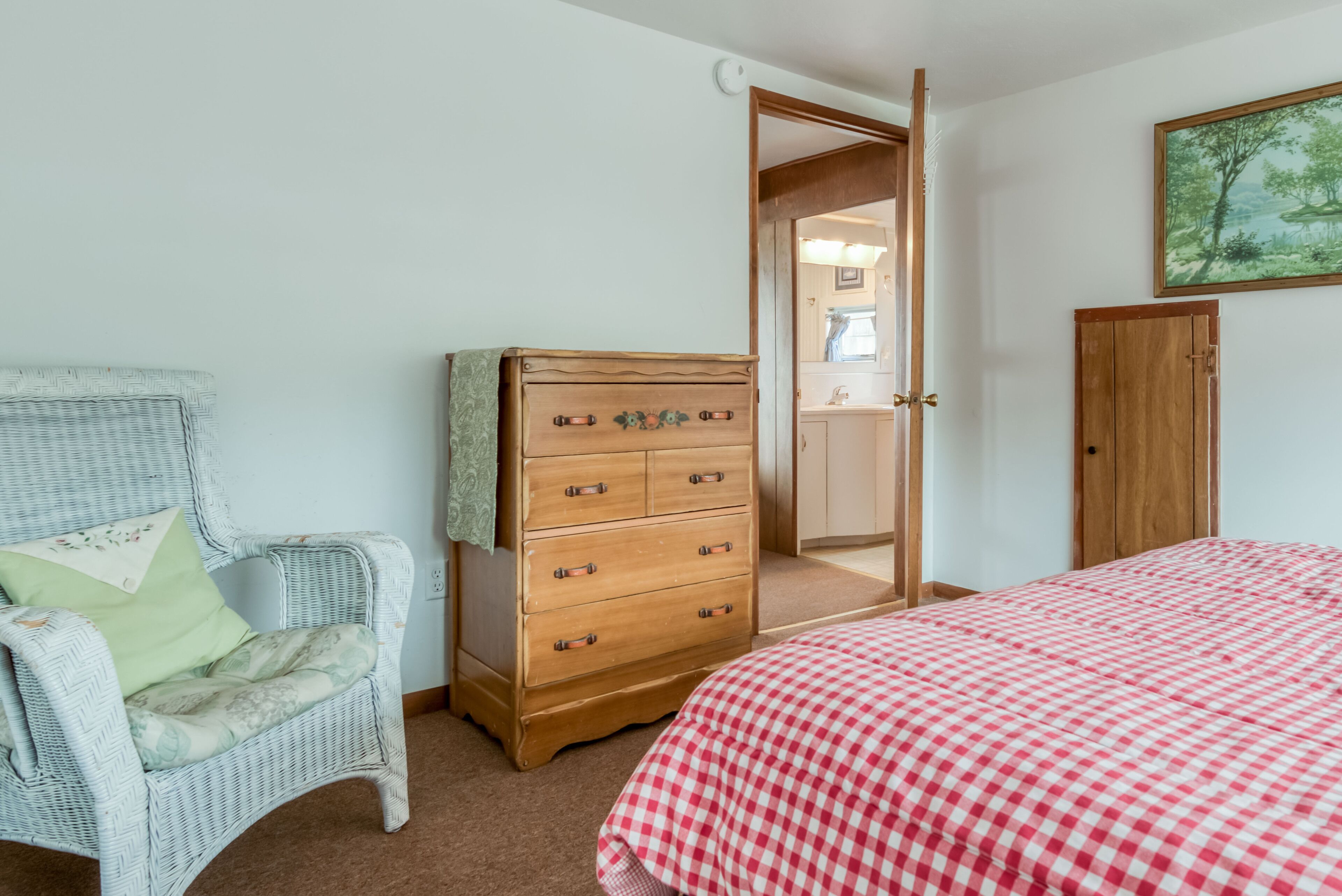 Een slaapkamer met een rieten stoel, een houten dressoir, een bed met ruitjesdeken en een deur naar de badkamer.