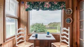 Ein Holzinnenraum mit zwei weißen Stühlen und einem Tisch, ein Fenster mit Blick auf den Strand und zwei an der Wand befestigte dekorative Teller.