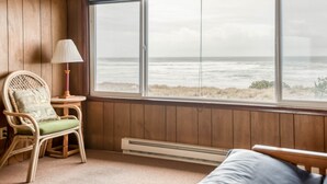 Ein Zimmer mit großem Fenster mit Blick aufs Meer, ein Korbstuhl mit Kissen und ein kleiner Tisch mit Lampe.