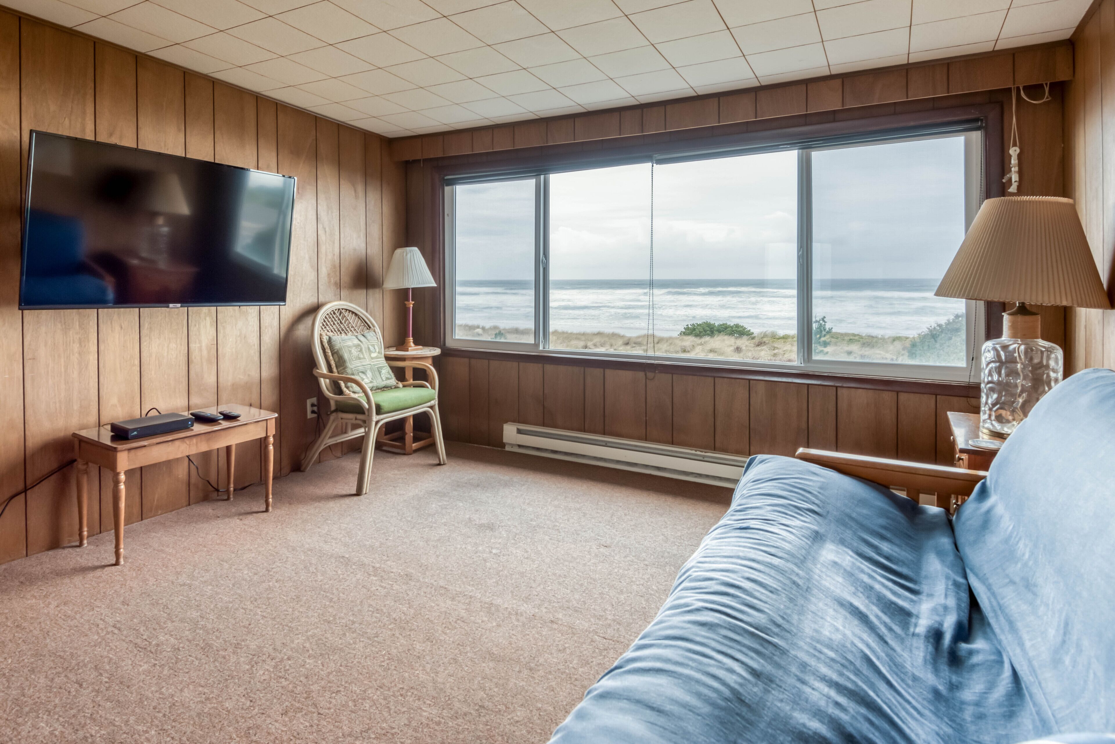 Una habitación con un ventanal grande con vista al océano, un televisor montado en la pared, piso de madera, un sofá azul y una silla con lámpara.