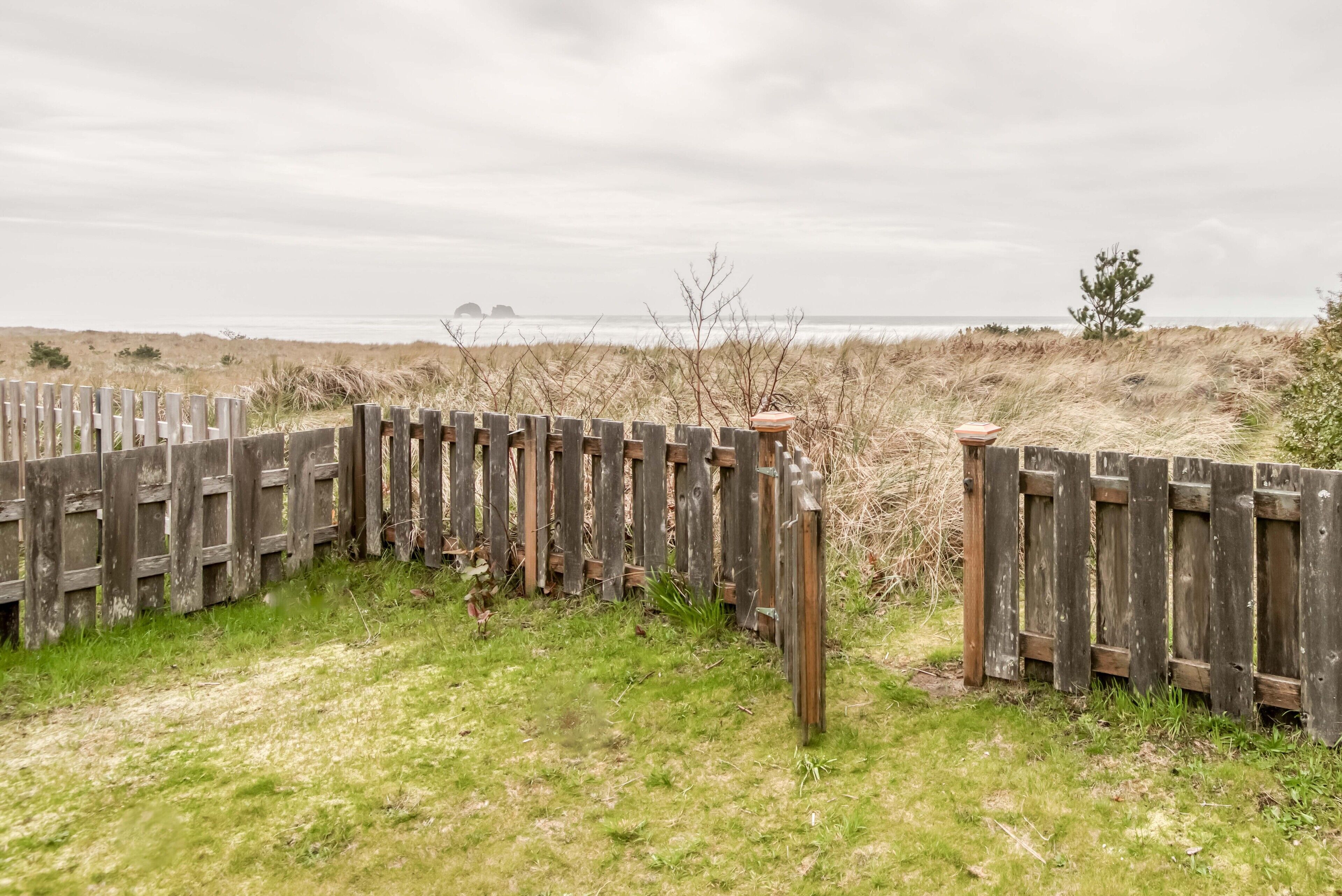 A wooden fence with a gate, a grassy area, and a view of the sea and dunes in the background.