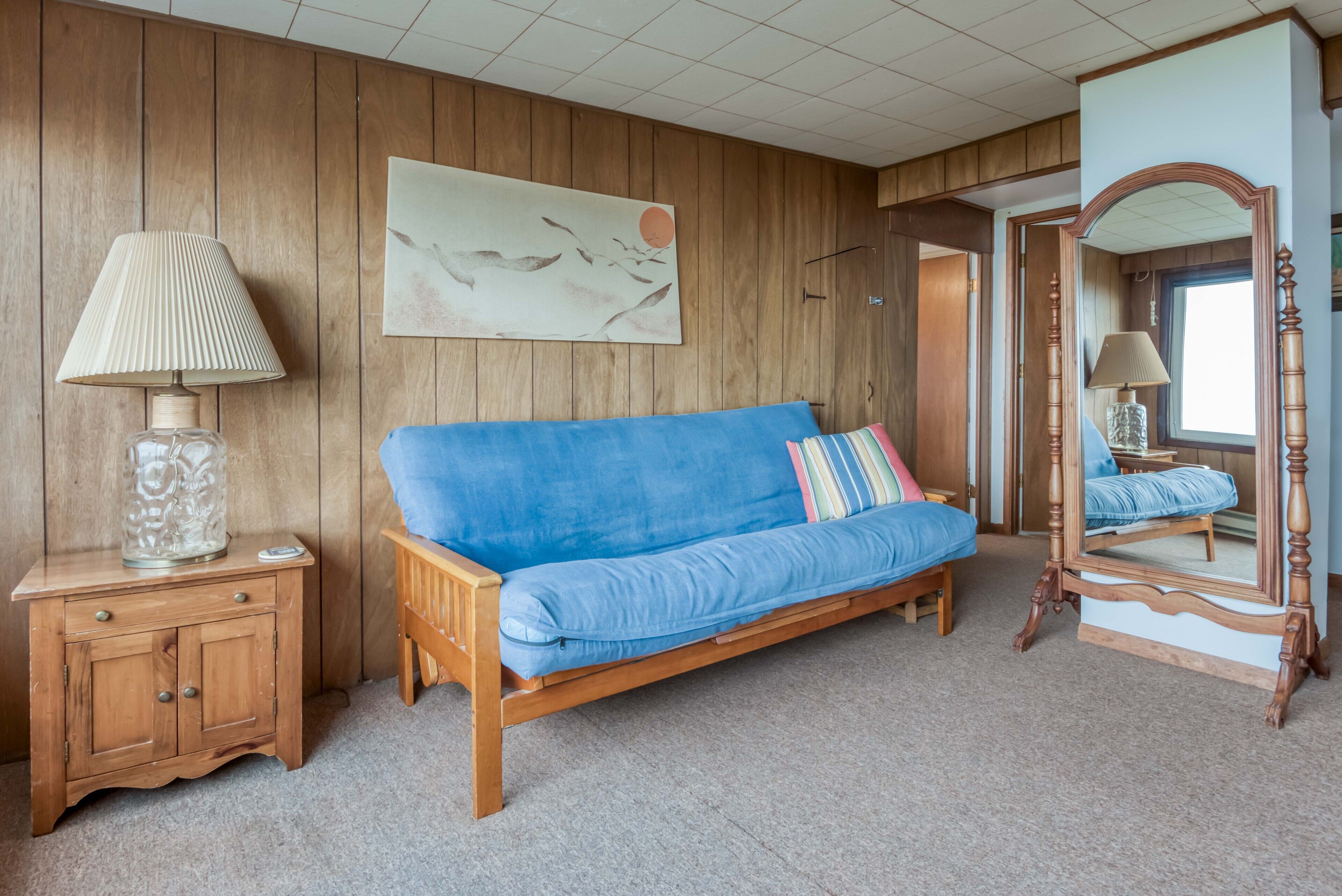 Quarto com parede de painel de madeira, sofá azul com almofada, mesa de cabeceira de madeira com abajur e espelho com moldura de madeira.