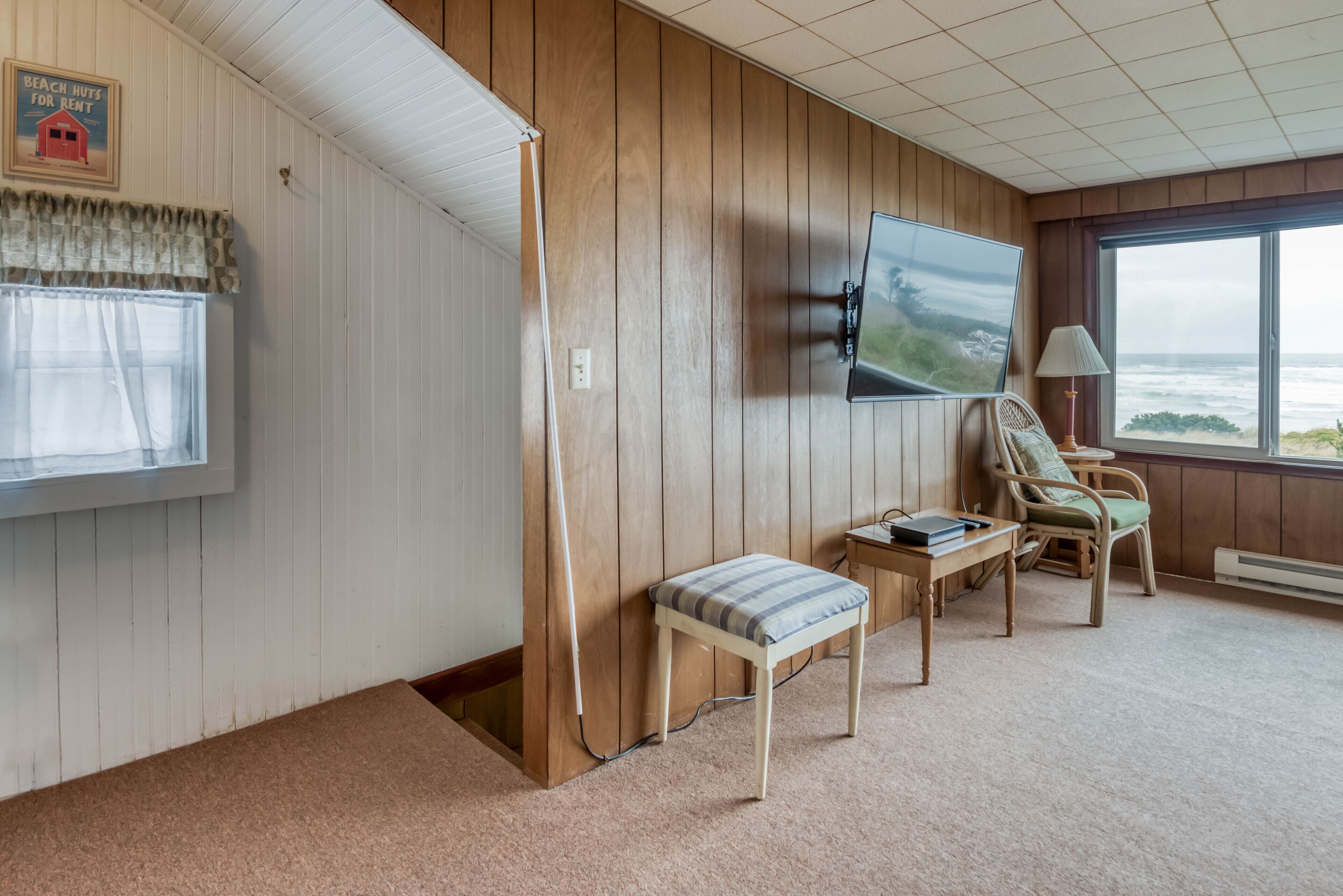 Una habitación con televisión montada en la pared, ventana con vista al océano, una banca y una silla.