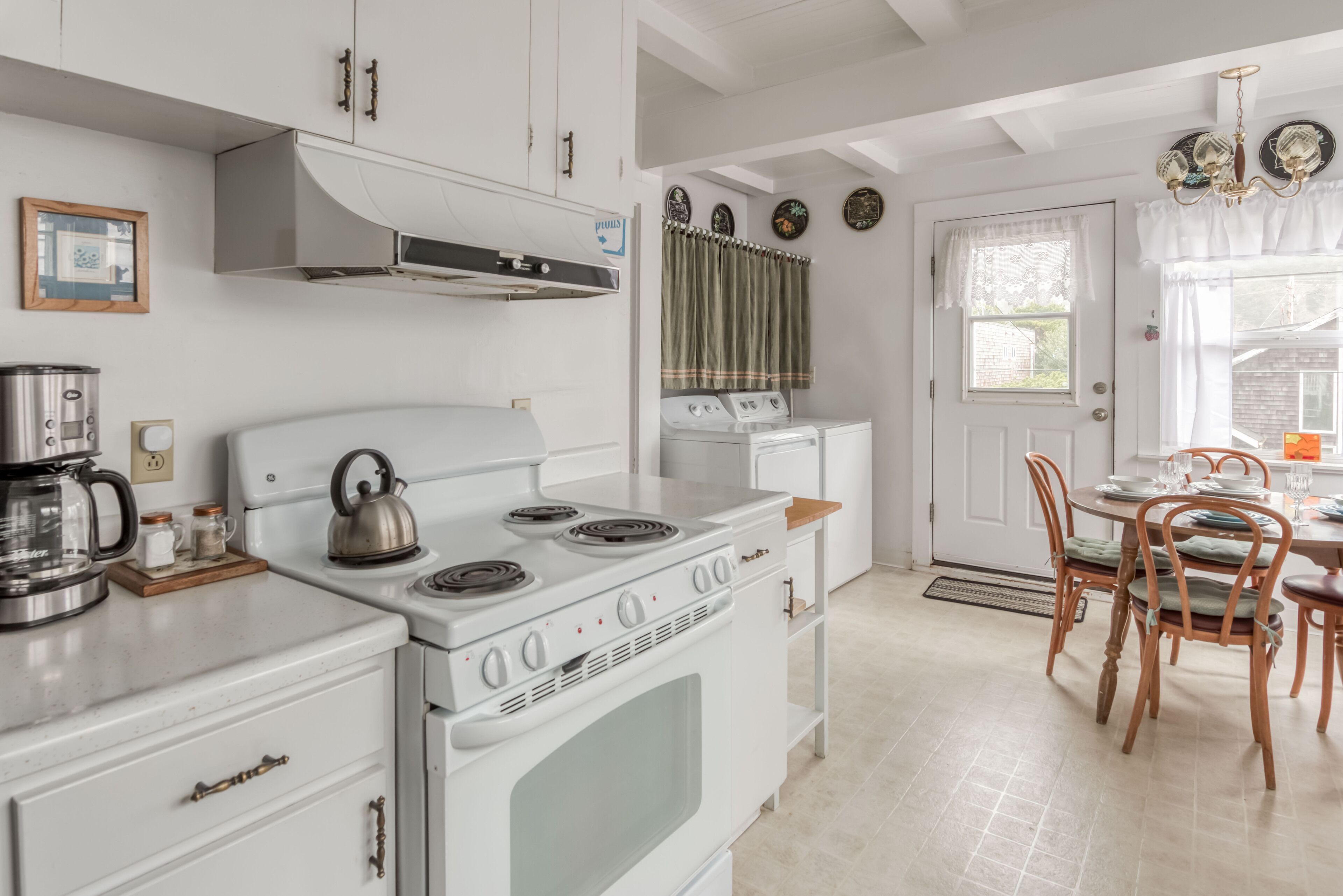 A kitchen with white appliances, a kettle, and a coffee maker. There is a dining table with chairs and a chandelier.