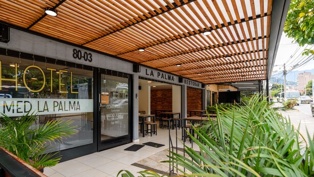 Façade d’un hôtel avec une pergola en bois, des sièges en plein air et une enseigne indiquant « HÔTEL MED LA PALMA ».
