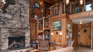 Een gezellig interieur van een lodge met een stenen open haard, houten wanden, een trap en een verzameling boeken en decoratieve objecten.