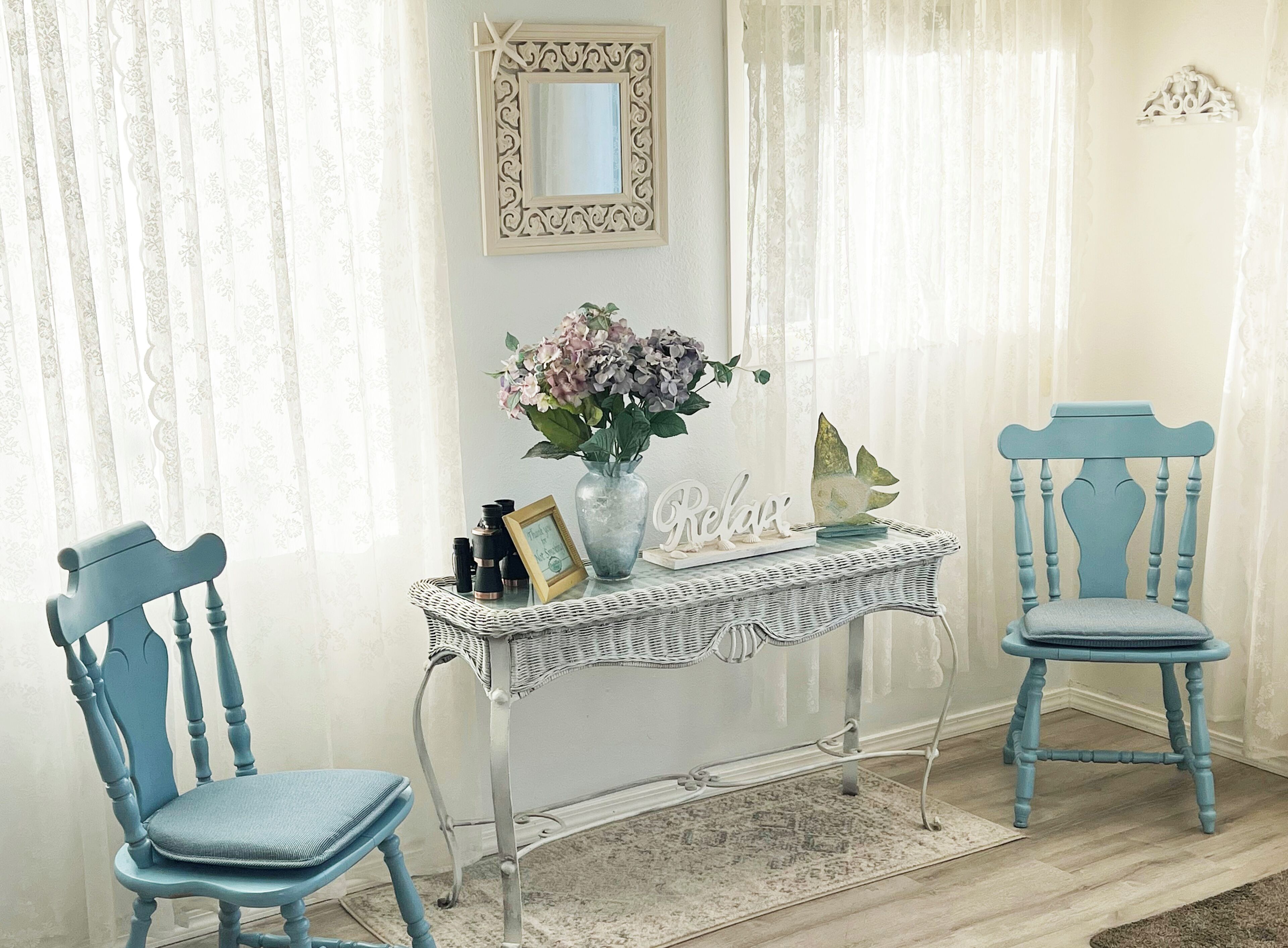 A room with two blue chairs, a white wicker table with a vase of flowers and a 'Relax' sign, a framed picture, and a decorative mirror on the wall.