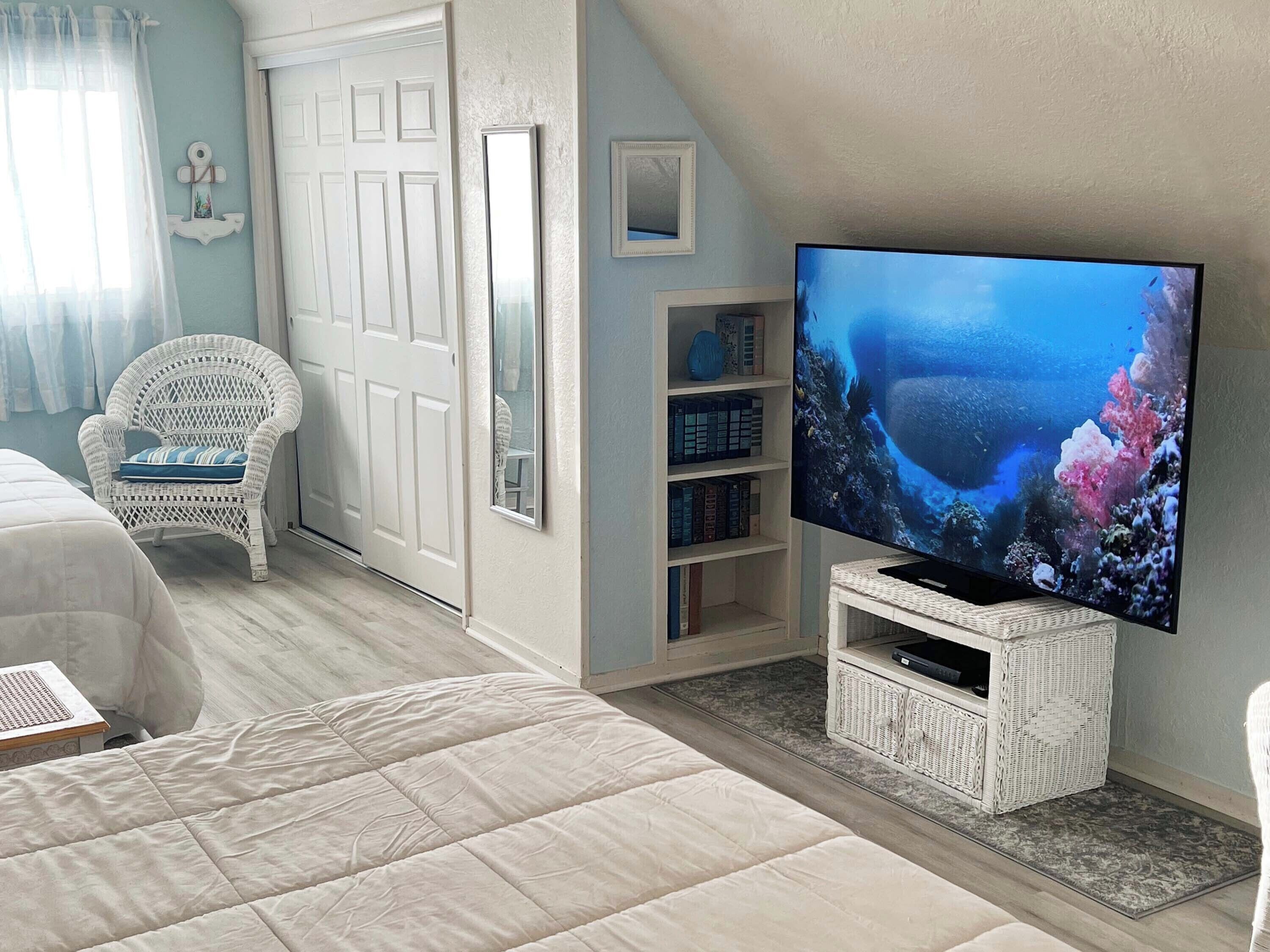 Un dormitorio con una cama grande, una silla de mimbre blanca, un televisor sobre un pedestal blanco y una estantería.