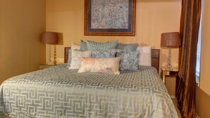 A bedroom with a large bed, patterned bedding, two bedside tables with lamps, and a framed artwork above the bed.