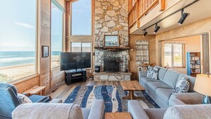 A spacious living room with a stone fireplace, large windows overlooking the beach, and a cozy seating area. - Oceanfront Home, Deck, High Windows with Amazing Views, Beach Access Nearby (Lincoln City)