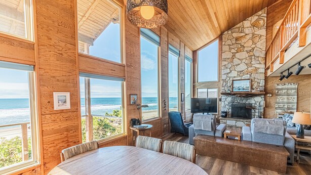 A spacious living room with a large wooden table, comfortable seating, a stone fireplace, and a view of the ocean. - Oceanfront Home, Deck, High Windows with Amazing Views, Beach Access Nearby (Lincoln City)