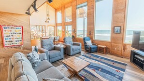 A spacious living room with a large sofa, armchairs, a coffee table, and a TV. The room has large windows offering a view of the ocean. - Oceanfront Home, Deck, High Windows with Amazing Views, Beach Access Nearby (Lincoln City)