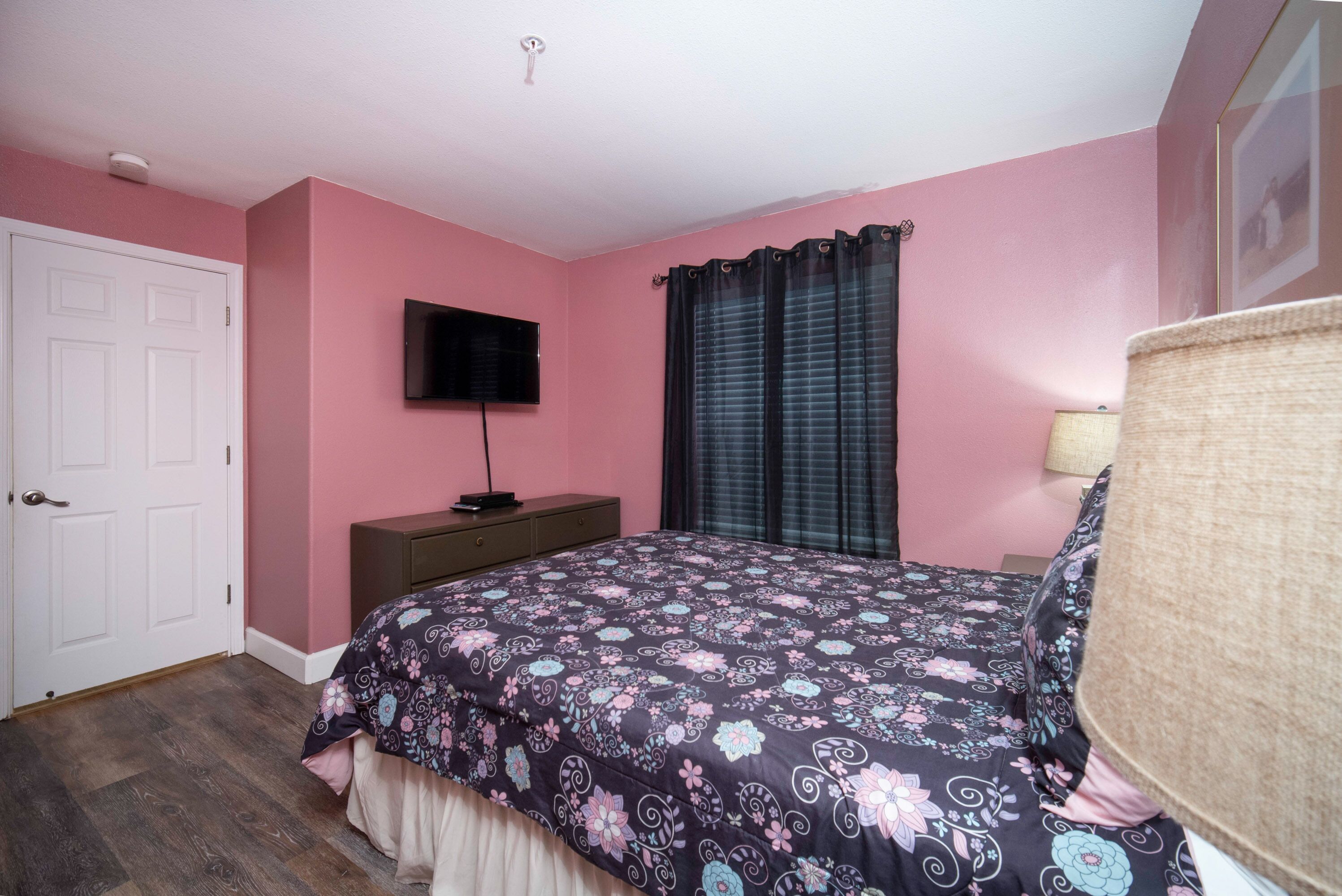 Slaapkamer met roze wanden, een bed met bloemenprint, een televisie aan de muur, een donkere houten dressoir en een lamp op het nachtkastje.