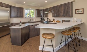 Cozinha moderna com armários de madeira escura, geladeira de aço inoxidável, uma ilha grande com banquetas e vista para o exterior pela janela.