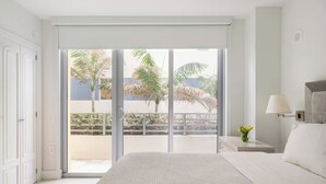 A bedroom with a large bed, a nightstand with a lamp, and a view of a balcony with palm trees. - Private Condos at 101 Ocean by LMC (Miami Beach)