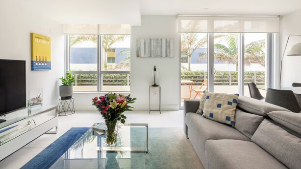 A modern living room with a sofa, a coffee table, a TV, and a large window with a view of palm trees and buildings. - Private Condos at 101 Ocean by LMC (Miami Beach)