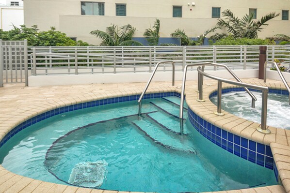 Une piscine dont le bord est revêtu de carreaux, dotée d’une rambarde métallique et offrant une vue sur un bâtiment avec des fenêtres et de la verdure en arrière-plan.