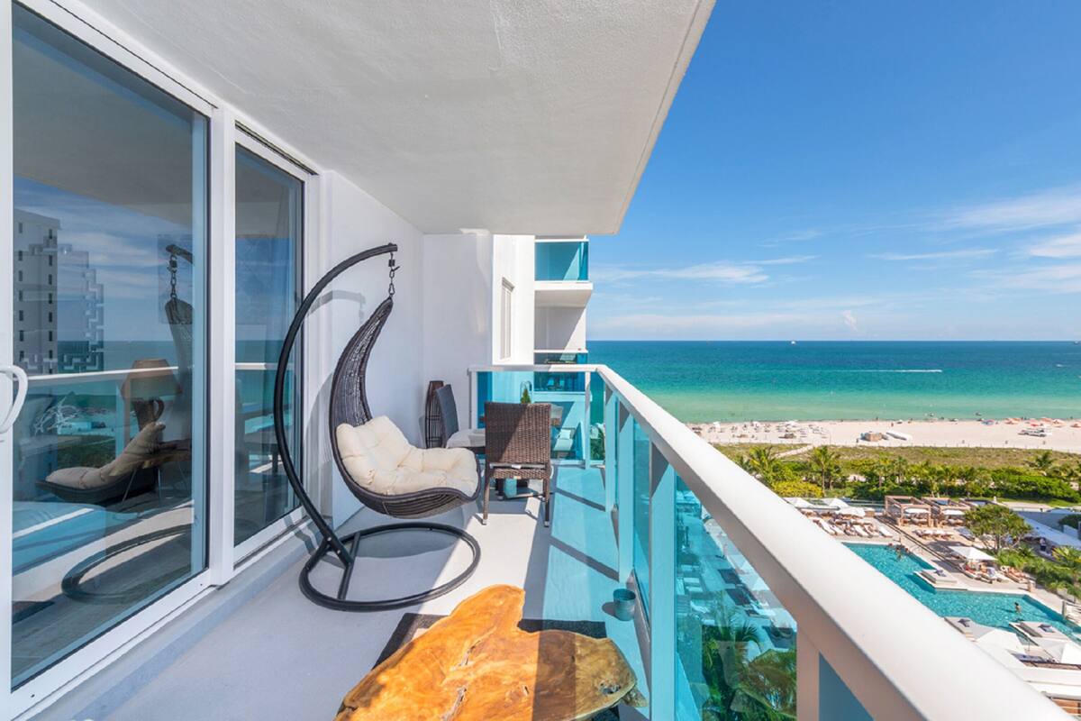 A balcony with a view of the ocean, a lounge chair, a dining table, and a pool area.