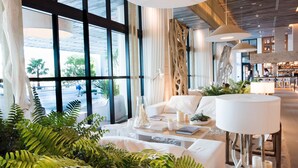A modern hotel lobby with large windows, a seating area with a white sofa and a coffee table, and a bar area in the background. - 1 Homes Vacation Rentals by LMC (Miami Beach)