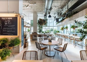 Um café moderno com mesas redondas e cadeiras de vime, rodeado por plantas em vasos e grandes janelas.
