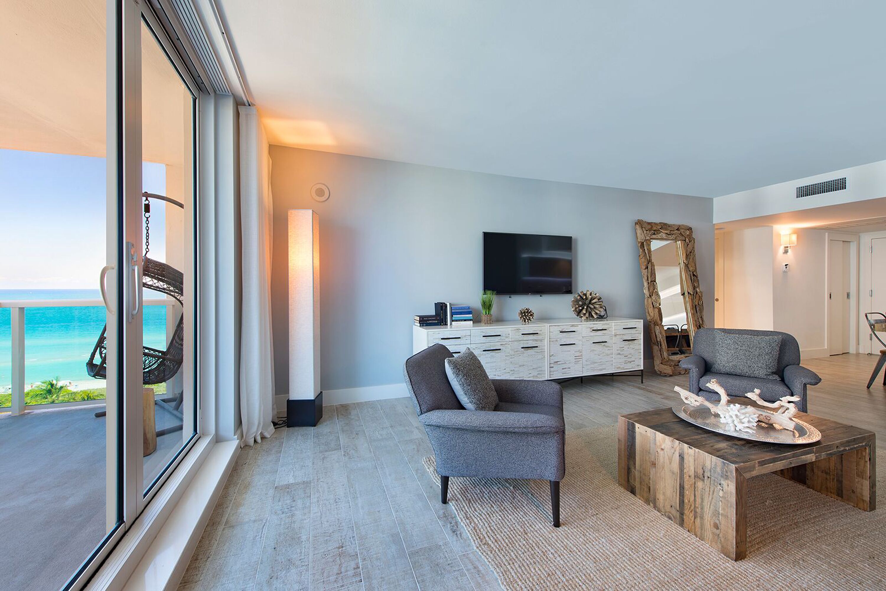 A modern living room with a view of the ocean, a large window, a flat-screen TV, a grey armchair, a wooden coffee table, and a white cabinet with decorative items.