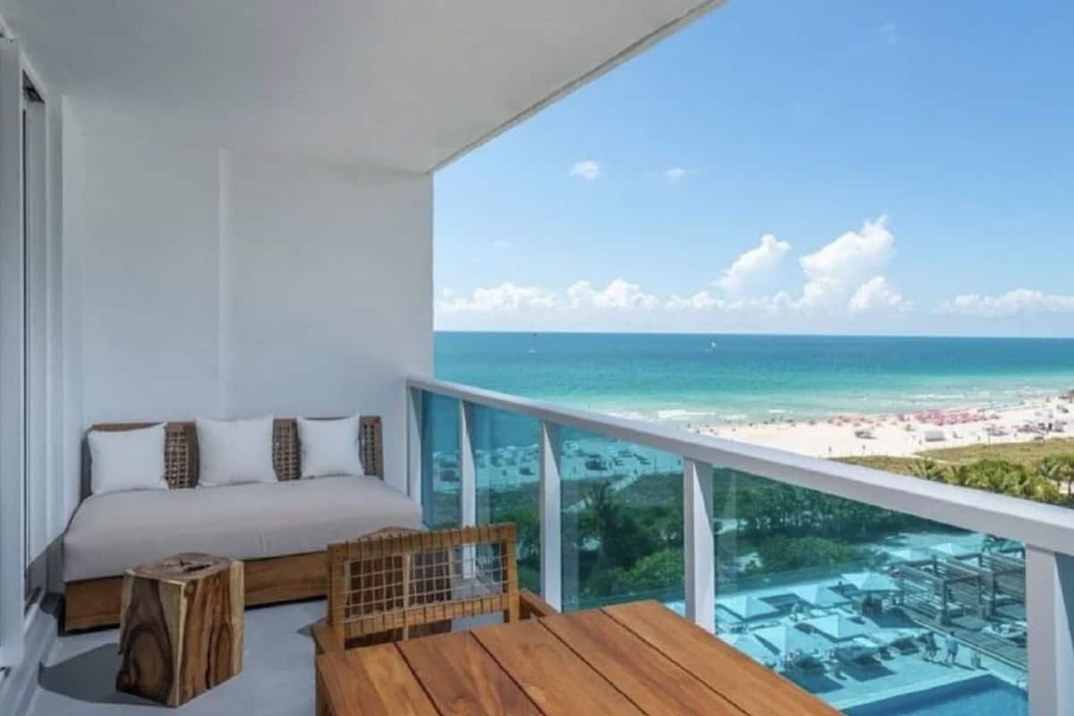 A balcony with a bed, wooden furniture, and a view of the beach and ocean.