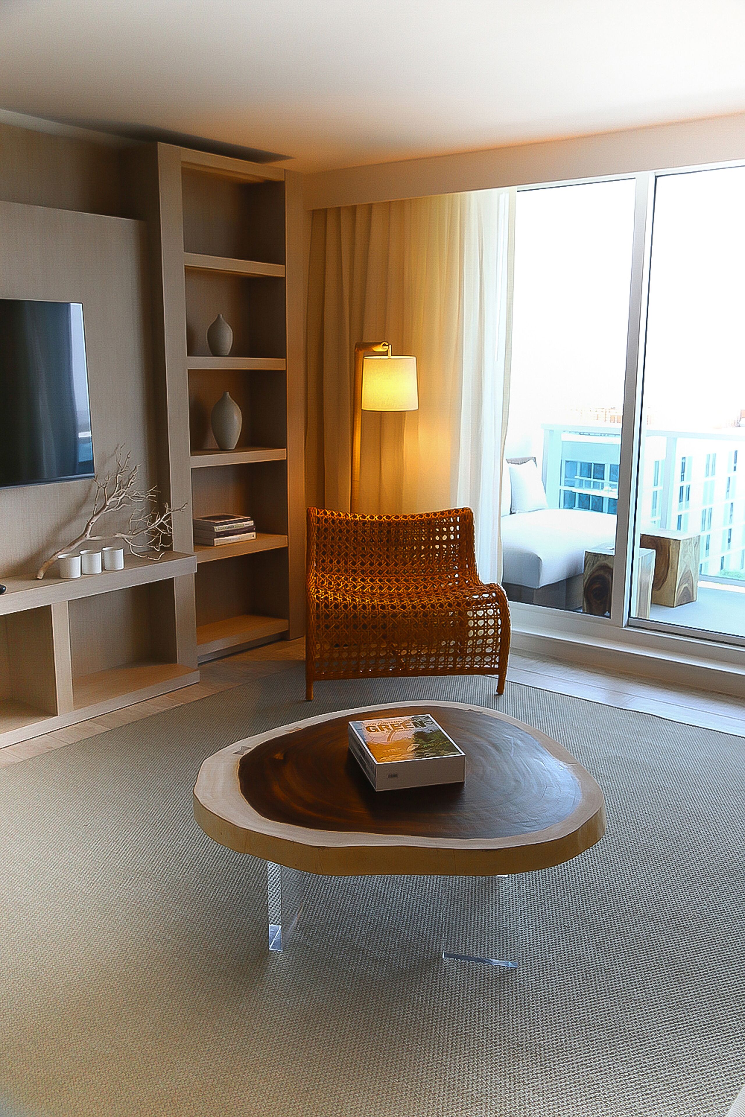 A hotel room with a large window, a wicker chair, a round coffee table, and a TV mounted on the wall.